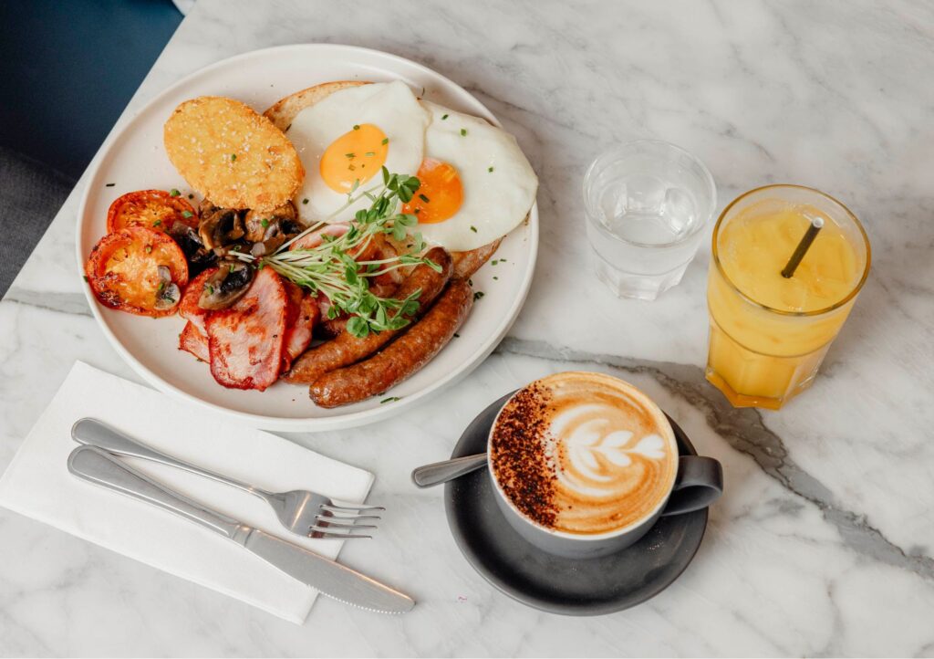 Hearty breakfast at Outside In Kitchen, Diggers The Entrance, featuring eggs, bacon, sausages, mushrooms, grilled tomato, hash brown, barista-made coffee and fresh juice served on a marble table.
