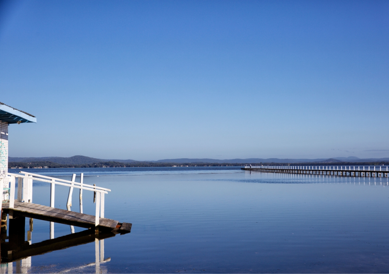 Calm morning view of Tuggerah Lake with a long timber jetty, glassy water and clear blue skies on the NSW Central Coast near The Entrance.
