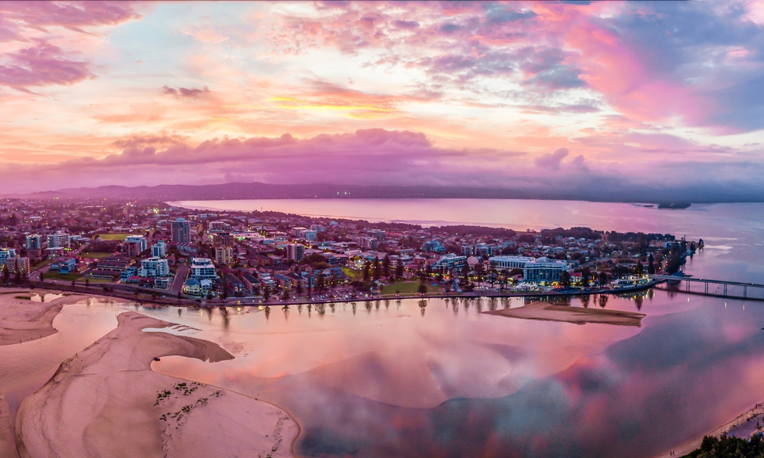 Aerial view of The Entrance at sunset, with pink skies reflected across Tuggerah Lake and the waterways of the NSW Central Coast.