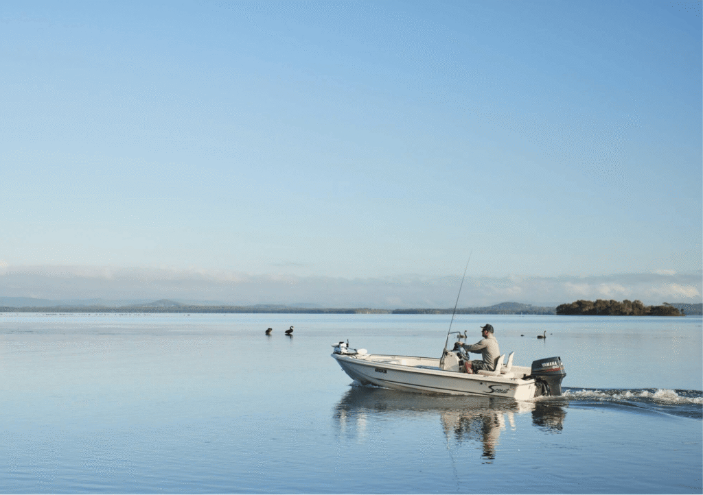 A man travelling in a small motorboat with fishing rods across the calm waters of Tuggerah Lakes near The Entrance, NSW, with black swans, distant hills and a clear blue sky.